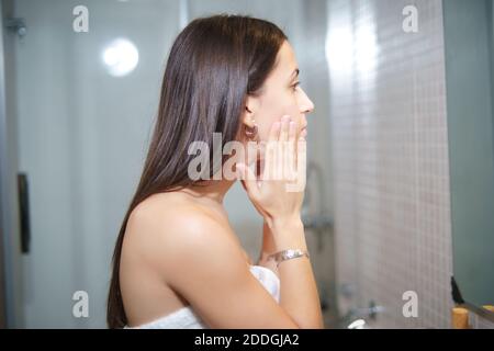 Seitenansicht der ruhigen Frau mit perfekter Haut Anwendung Gesichtsbehandlung Creme während der täglichen Schönheitsbehandlung im Badezimmer Stockfoto