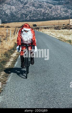 Weihnachtsmann im roten Kostüm Reiten moderne Fahrrad entlang leer Straße im Hochland an sonnigen Tag Stockfoto