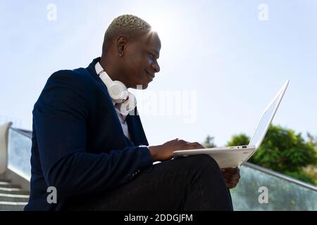 Seitenansicht des Inhalts African American männlich Unternehmer in stilvoll Tragen Sie sitzen auf Treppen im Stadtzentrum und tippen Sie auf Laptop während der Arbeit aus der Ferne o Stockfoto