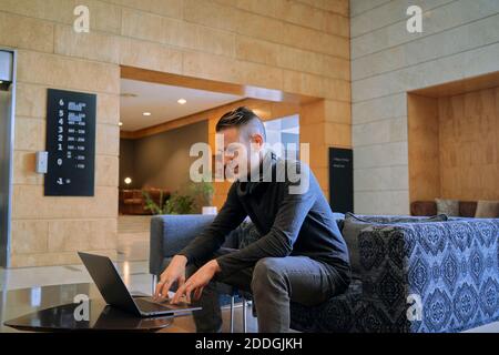 Seitenansicht des konzentrierten männlichen Unternehmers, der in einem Sessel sitzt Lounge des Hotels, während man einen Laptop benutzt und an einem Remote-Projekt arbeitet Stockfoto