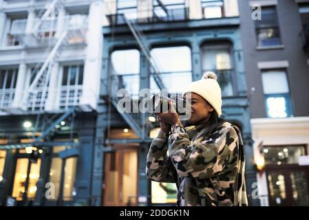 Niedriger Winkel der afroamerikanischen Fotografin in warmen Hut Und stylischer Pullover, der moderne Fotokameras fotografiert New York City Stockfoto