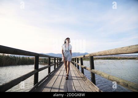 Ganzer Körper von zuversichtlich junge Frau in lässigen Sommerkleid Spaziergang auf hölzernen Steg über dem See in sonnigen Sommertag Stockfoto