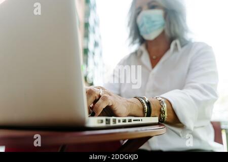 Niedriger Winkel der mittleren Alters weibliche Freiberufler in Schutzmaske Sitzen am Tisch im Café im Freien und arbeiten aus der Ferne Projizieren, während Netbook verwendet wird Stockfoto