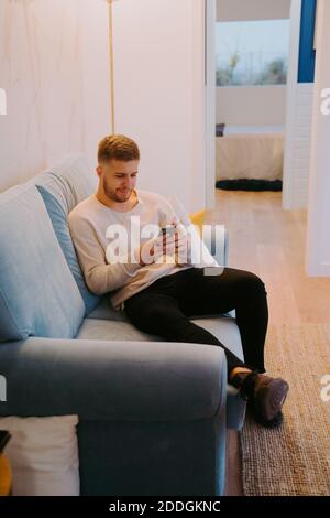 Entspannter junger Mann, der auf dem Sofa chillt und eine SMS schreibt Smartphone, während Sie das Wochenende zu Hause verbringen Stockfoto