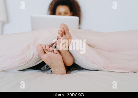 Junge ethnische barfuß Weibchen mit Decke im Bett sitzend bedeckt Und das Surfen im Laptop zu Hause Stockfoto