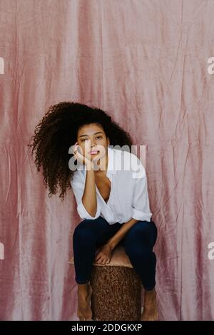 Selbstbewusste Millennial ethnischen Weibchen mit Afro lockiges Haar tragen weiß Bluse und Jeans sitzen gegen rosa Vorhang Blick auf die Kamera Stockfoto