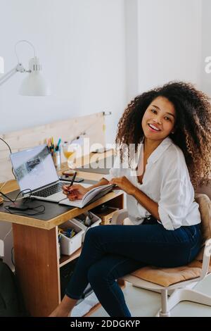 Seitenansicht der modernen jungen glücklichen afroamerikanischen Frau fernab Mitarbeiter in lässiger Kleidung sitzt am Tisch mit Laptop und Schreiben in Planer während Wor Stockfoto