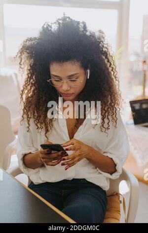 Serious afroamerikanische Unternehmerin mit Abuds sitzen am Tisch Im Heimbüro und beim Lesen von Nachrichten auf dem Handy während der Ferne Arbeiten Stockfoto