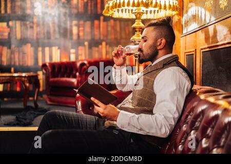 Seitenansicht des entspannten bärtigen Mannes mit Tablet und genießen Alkohol, während Sie sich auf dem Sofa in einem eleganten Pub ausruhen Stockfoto