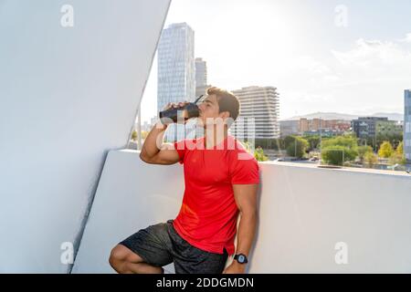 Junge muskulösen männlichen in Sportbekleidung Trinkwasser aus Plastikflasche Während Sie sich nach dem intensiven Training auf der Terrasse der modernen Stadt ausruhen Gebäude Stockfoto