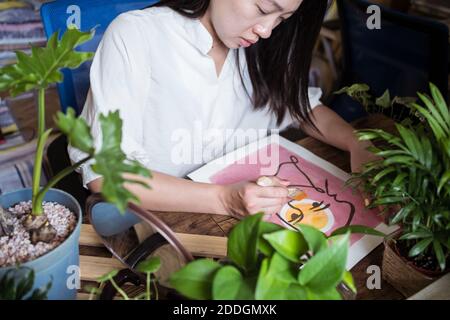 Fokussierte asiatische Künstlerin sitzen am Holztisch und schaffen Bild auf Papier mit Farbe und Pinsel Stockfoto