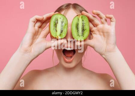 Porträt eines fröhlichen weiblichen Mädchen, natürliche klare Haut, ein Mädchen mit zwei Stücken von Kiwi, bedeckt ihre Augen, isoliert auf einem rosa Hintergrund. Hautpflege Stockfoto