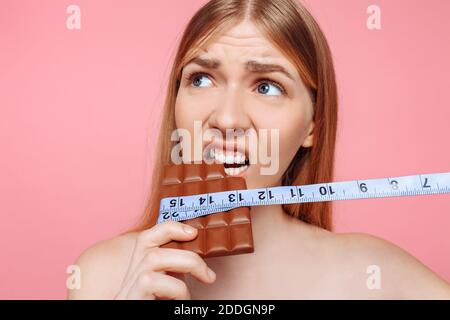 Portrait eines nachdenklichen Mädchens beißt eine Tafel Schokolade mit einem Maßband auf einem rosa Hintergrund gewickelt. Ernährungskonzept Stockfoto
