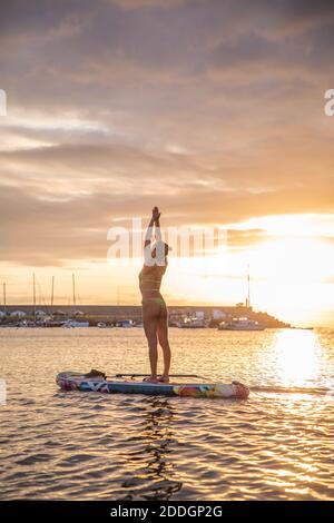 Seitenansicht Silhouette der schlanken weiblichen stehend in Berg Pose Mit den Armen nach oben beim Yoga auf dem schwimmenden Paddle Board Gegen den Sonnenuntergang Stockfoto