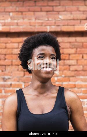 Junge glückliche afroamerikanische Frau mit lockigen Haaren, die dagegen stehen Ziegelmauer und nachdenklich weg schauen Stockfoto