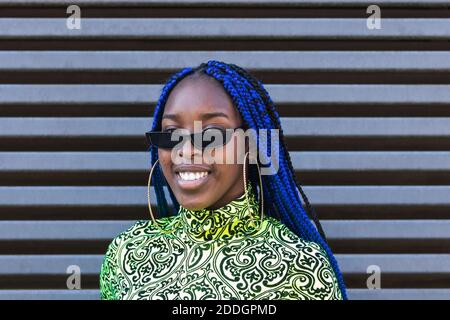 Selbstbewusst glücklich afroamerikanische Frau mit blauen Zöpfen tragen trendy Outfit und Sonnenbrille stehen auf der Straße und schauen auf die Kamera Stockfoto