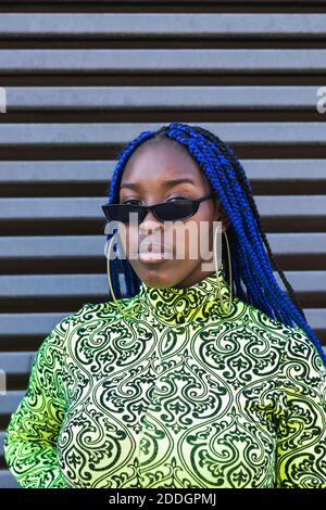 Selbstbewusste afroamerikanische Frau mit blauen Zöpfen tragen trendige Outfit Und Sonnenbrillen stehen auf der Straße und schauen auf die Kamera Stockfoto