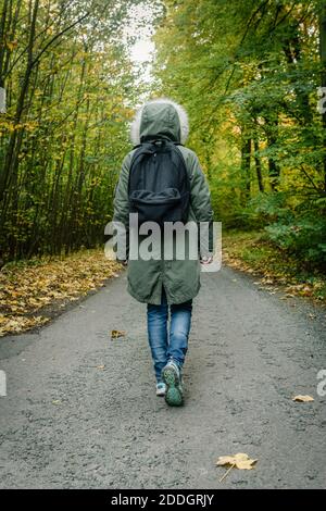 Rückansicht von Explorer in Oberbekleidung und mit Rucksack zu Fuß Entlang der Straße in grünen Wäldern während des Abenteuers Stockfoto
