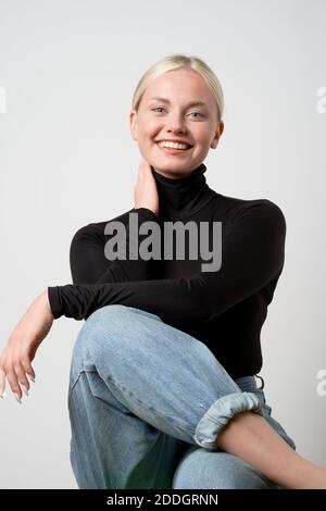 Fröhliche blonde Frau in lässige Kleidung sitzt im Studio und Lachen Sie aufrichtig, während Sie die Kamera betrachten Stockfoto