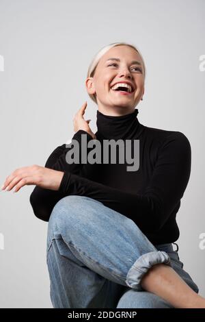 Fröhliche blonde Frau in lässige Kleidung sitzt im Studio und Lachen Sie aufrichtig, während Sie wegschauen Stockfoto