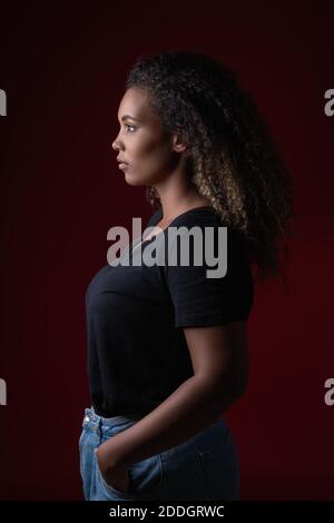 Seitenansicht einer jungen übergewichtigen afroamerikanischen Frau in einem lässigen schwarzen Hemd und Jeans, die im roten Studio stehen und wegschauen Stockfoto