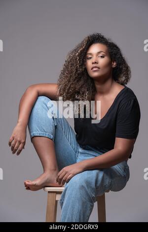 Plus Größe barfuß junge afroamerikanische Frau mit lockigen Haaren Gekleidet in Jeans und schwarzes Hemd sitzen auf Stuhl und Blick auf die Kamera gegen Grau Stockfoto