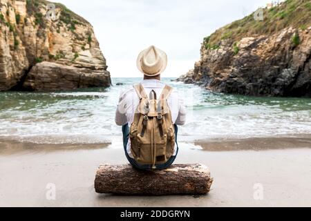 Rückansicht des nicht erkennbaren männlichen Rucksacktouristen im Hut sitzend auf Holzbalken an der Küste neben dem Meer mit felsigen Klippen und Entspannung während der Sommerreise Stockfoto