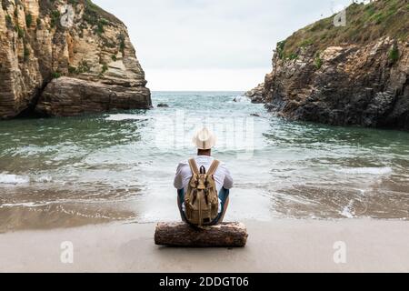 Rückansicht des nicht erkennbaren männlichen Rucksacktouristen im Hut sitzend auf Holzbalken an der Küste neben dem Meer mit felsigen Klippen und Entspannung während der Sommerreise Stockfoto