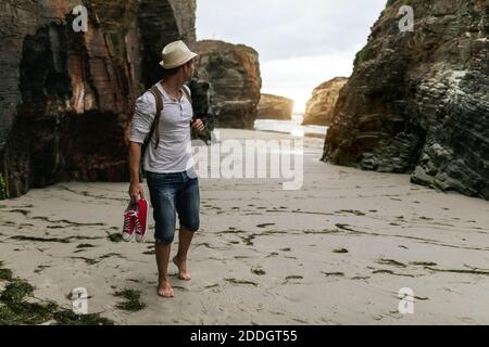 Barfuß männlicher Rucksacktourist mit Sneakers in der Hand, der alleine unterwegs ist Sandstrand in der Nähe des Meeres und bewundern Sie den Sonnenuntergang, während Sie sich danach entspannen Wandern in der Natur Stockfoto