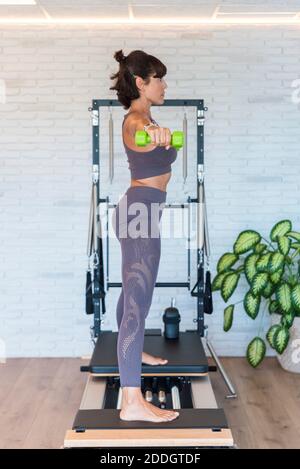 Seitenansicht der barfuß-Hündin, die die Arme ausstreckt, mit Hanteln Während des Pilates-Trainings im Fitnessstudio auf dem Reformer stehen Stockfoto