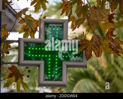 Grünes Kreuz Apotheke beleuchtete Schild hängend außerhalb Drogerie, Baum Laub Hintergrund Stockfoto