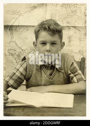 Originalfoto aus den 50er Jahren von einem Schuljungen, der ein kariertes Hemd und ein Trägerhemd trägt, sitzt am Schreibtisch und liest ein Buch, eine Karte an der Wand hinter ihm, Großbritannien Stockfoto