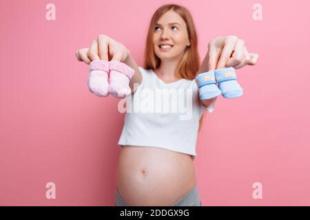 Junge oder Mädchen schöne schwangere Frau hält blau und rosa Baby Booties, posiert im Studio auf einem rosa Hintergrund Stockfoto