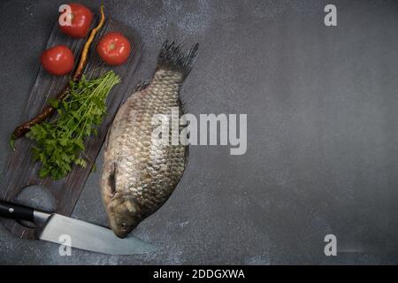 Roher Fischkarpfen vor dem Schneiden mit einem Holzbrett und Tomaten und Grüns auf dunklem Hintergrund. Stockfoto