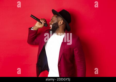 Leidenschaftliche schwarze männliche Sängerin vor rotem Hintergrund, singen ins Mikrofon, tragen Party-Outfit, stehen über rotem Hintergrund Stockfoto