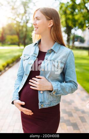 Glücklich schwangere Mädchen geht im Park, umarmt ihren Bauch, Stockfoto