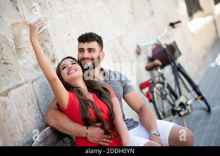 Pärchen sitzt auf einer Bank und nimmt ein Selfie mit einem Handy, während im Urlaub. Stockfoto
