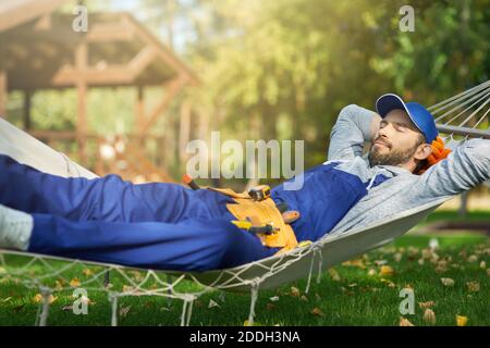 Entspannter junger männlicher Baumeister, der blaue Overalls und Kappe trägt, die eine Pause machen, liegend in einer Hängematte draußen mit geschlossenen Augen an einem sonnigen Tag. Gebäude, Beruf, Rest-Konzept Stockfoto