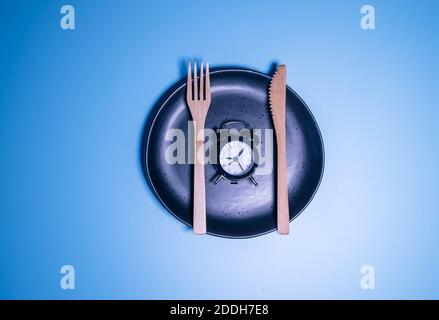 Wecker und ein Teller mit Besteck isoliert auf blauem Hintergrund. Konzept der intermittierenden Fasten, Mittagessen, Ernährung und Gewichtsverlust. Stockfoto