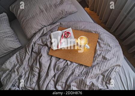 Blick von oben auf die gemütliche Schlafzimmer mit großem Bett, in beigen Farben, Kaffeetasse, Peppermint Candy Cane und Buch. Faul Weihnachten Morgen im Bett Stockfoto