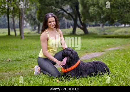 Black briard liegt mit der Zunge draußen und Frau in der Nähe im öffentlichen Park. Hund mit langen Haaren liegt auf dem Gras im Freien. Stockfoto