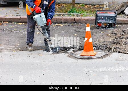 Ein Straßenarbeiter in reflektierender Kleidung zerschlägt alten Asphalt mit einem elektrischen Presshammer in der Nähe eines Schachtelschlochs. Stockfoto