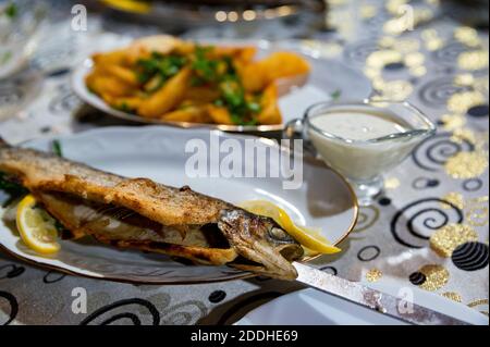 Drei leckere frittierte geröstete Fische mit Gewürzen auf einem Holzkohlesieb mit Zitrone, weißer Sauce und gebratenen Kartoffeln Stockfoto