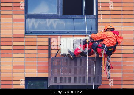 Ein Mann in roter Uniform verbindet die Klimaanlage Unter dem Fenster der Wohnung Stockfoto