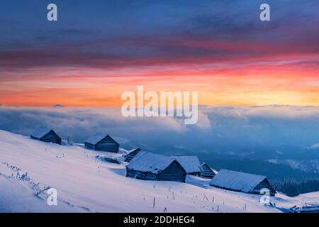 Fantastische Winterlandschaft mit Holzhaus in verschneiten Bergen während unglaublichen orangen Sonnenuntergang. Weihnachtsfeiertagskonzept Stockfoto