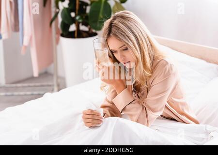 Frau in Schlafanzug hält Glas Wasser und Glas mit Pillen auf dem Bett Stockfoto