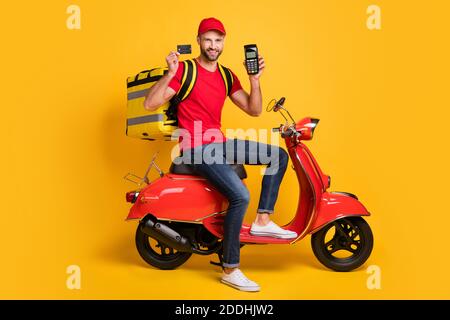 Foto von Mann Fahrt Roller halten Terminal Kreditkarte tragen Tasche rot T-Shirt Kappe isoliert gelbe Farbe Hintergrund Stockfoto