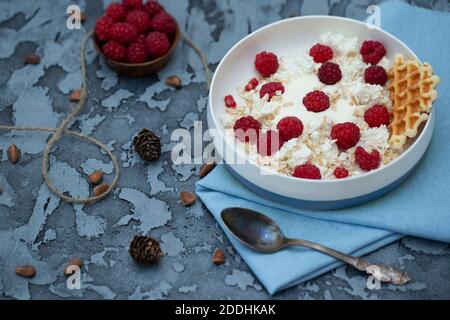 Hüttenkäse auf einem Teller mit Joghurt, bestreut mit Himbeeren und Müsli, blau auf einem Handtuch, Metalllöffel und Nüsse. Stockfoto