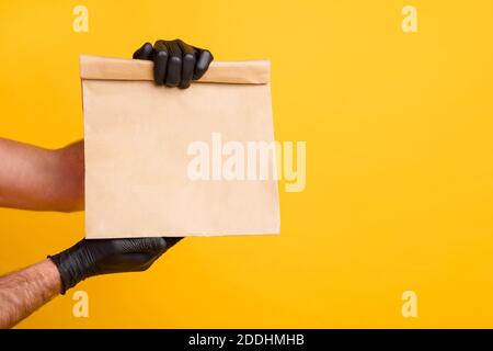 Zugeschnittenes Foto der Lieferung Mann Arme halten Takeout Papierpaket Tragen Sie schwarze Handschuhe isoliert leuchtend gelben Hintergrund Stockfoto