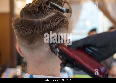 Ein Friseur schneidet einen bärtigen jungen Kerl mit einem Haarschneider und kämmt die Haare auf seinem Kopf. Die Arbeit des Meisters im Männerhaarschnitt in einem Friseurladen. Die Ba Stockfoto